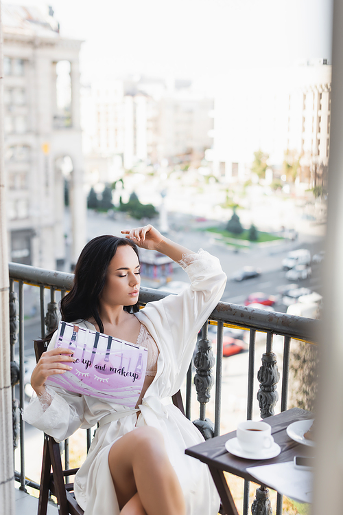 brunette woman in white robe sitting on balcony and reading magazine