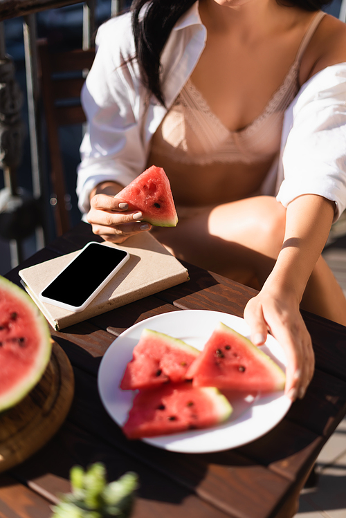 cropped view of woman in beige underwear and white shirt eating watermelon on balcony