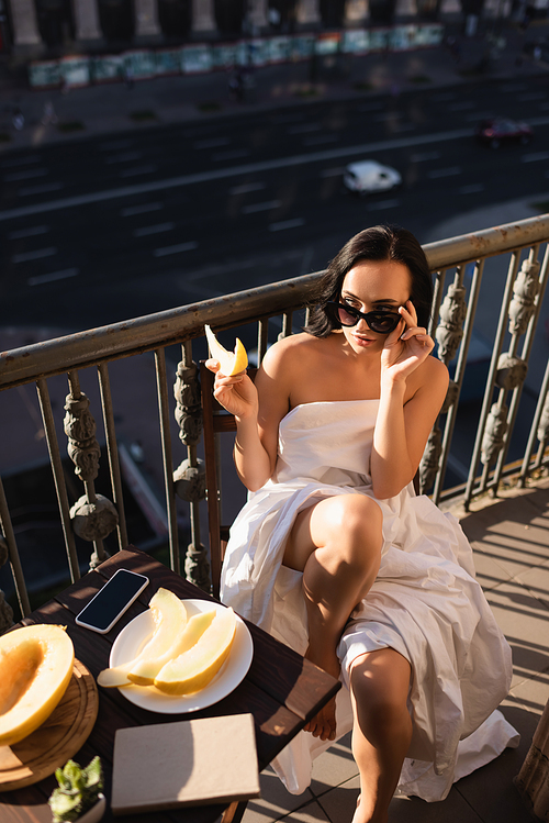 sexy brunette woman raising sunglasses and eating  on balcony