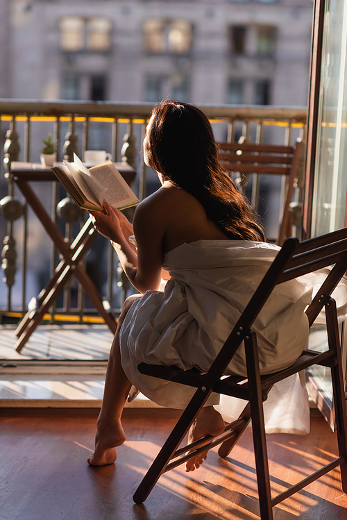 back view of attractive woman covered in white sheet reading book and sitting on balcony