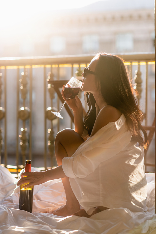 brunette woman in sunglasses and white shirt drinking wine on balcony