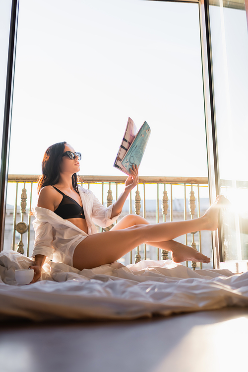 beautiful brunette woman in sunglasses and black underwear reading magazine and holding cup of tea