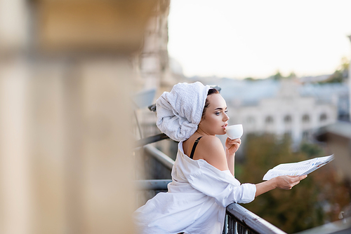 sexy young woman in white shirt with towel on head holding newspaper and drinking tea on balcony