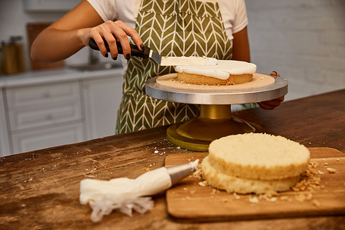 Cropped view of confectioner with spatula putting cream on sponge cake layer