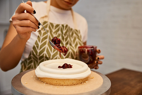 Cropped view of confectioner adding jam on sponge cake with cream