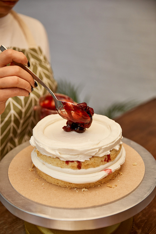 Cropped view of confectioner with spoon putting berry jam on cake with cream