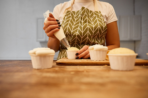 Selective focus of confectioner adding sweet cream on cupcakes on wooden table, cropped view