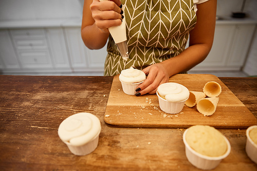 Cropped view of confectioner pouring cream on cupcakes beside waffle cones on cutting board