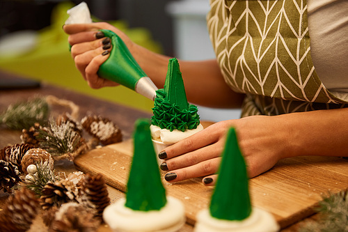 Selective focus of confectioner decorating tasty Christmas tree cupcakes beside spruce cones on table, cropped view