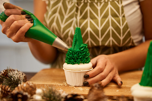 Cropped view of confectioner decorating Christmas tree cupcake with green cream on table with spruce cones