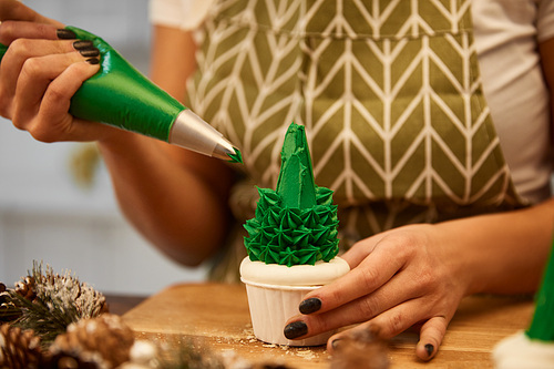 Cropped view of confectioner adding green cream on Christmas tree cupcake beside spruce cones on table