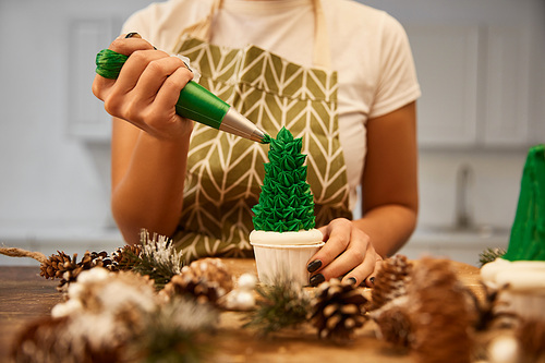 Cropped view of confectioner decorating Christmas tree cupcake with green cream beside spruce cones on table