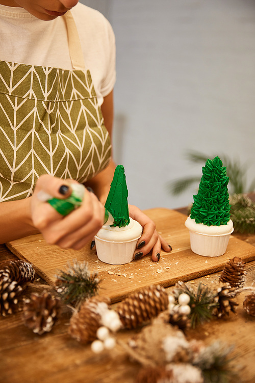 Cropped view of confectioner using pastry bag with cream on Christmas tree cupcakes beside spruce cones on table