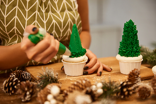 Selective focus of confectioner working with Christmas tree cupcakes beside spruce cones on table, cropped view