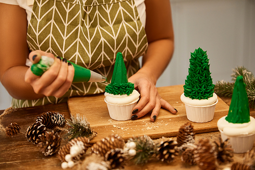 Cropped view of confectioner making Christmas tree cupcakes with green sweet cream beside spruce cones on table