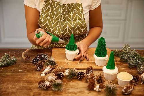 Cropped view of confectioner making Christmas tree cupcakes with cream beside spruce branches and cones on table