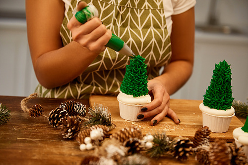 Cropped view of confectioner adding green sweet cream on Christmas tree cupcakes with spruce cones on table