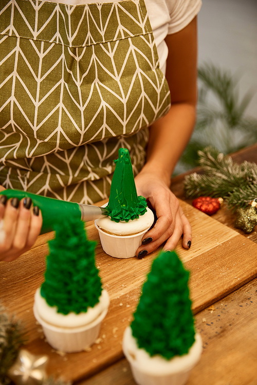 Cropped view of confectioner decorating Christmas tree cupcakes beside spruce branches and christmas balls on table