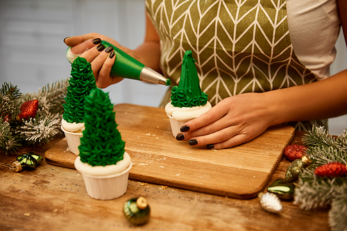 Cropped view of confectioner making Christmas tree cupcakes beside spruce branches and christmas balls on table