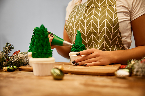 Cropped view of confectioner decorating Christmas tree cupcakes beside pine branch and christmas balls on table
