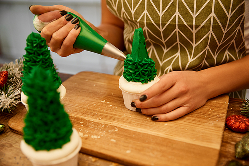 Cropped view of confectioner adding sweet cream on Christmas tree cupcakes with pine branches and christmas balls on table