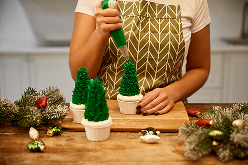 Cropped view of confectioner with pastry bag adding cream on Christmas tree cupcakes with pine branches on table