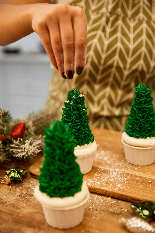 Cropped view of confectioner sprinkling decor on Christmas tree cupcakes beside christmas balls and spruce branch on table