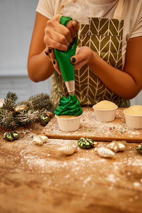 Cropped view of confectioner making cupcakes with green cream beside christmas decoration and pine branch on table