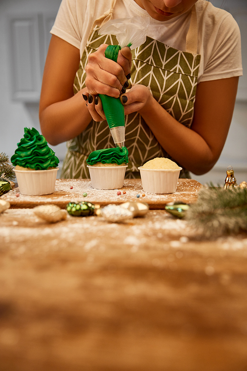 Cropped view of confectioner working with green cream and cupcakes beside baubles and pine branches on table