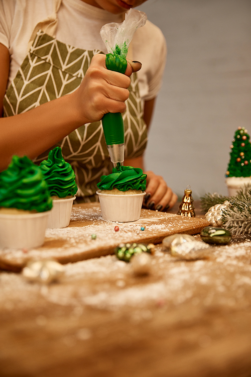 Cropped view of confectioner working with cream and cupcakes beside baubles and pine branch on table