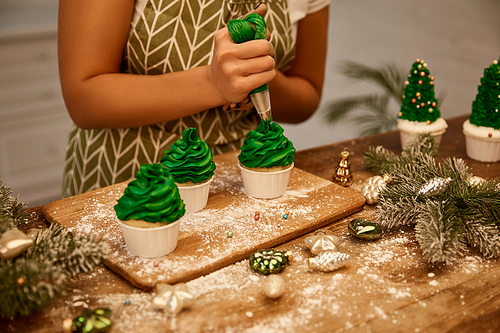 Cropped view of confectioner decorating Christmas tree cupcakes beside spruce branches and Christmas decorations on table