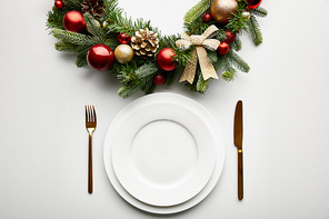 top view of white plates with golden cutlery near festive Christmas wreath with baubles on white background