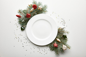 top view of festive Christmas table setting on white background with decorated pine branch, plate and confetti