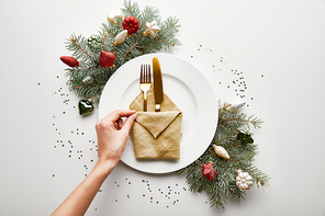 cropped view of woman holding napkin on white plate with cutlery near festive Christmas tree branch and confetti on white background