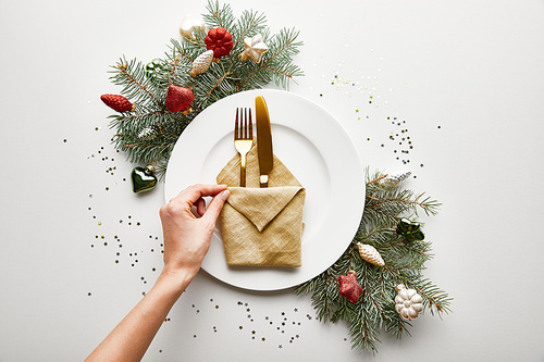 cropped view of woman holding napkin on white plate with cutlery near festive Christmas tree branch and confetti on white background