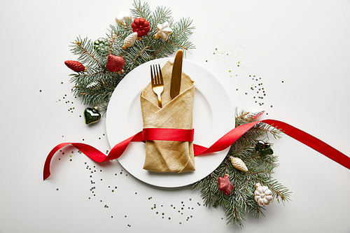 top view of white plate with napkin, cutlery and ribbon near festive Christmas tree branch with baubles and confetti on white background
