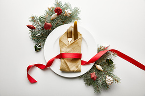 top view of white plate with napkin, cutlery and ribbon near festive Christmas tree branch with baubles on white background