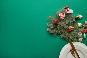 top view of white plate with cutlery near festive Christmas tree branch with baubles on green background with copy space