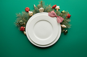 top view of white plates near festive Christmas tree branch with baubles on green background