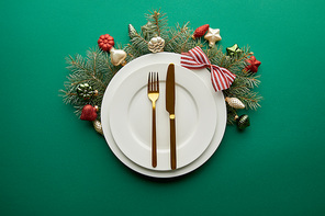 top view of white plates with cutlery near festive Christmas tree branch with baubles on green background