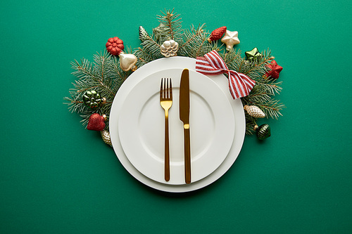 top view of white plates with cutlery near festive Christmas tree branch with baubles on green background