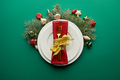 top view of white plate with napkin, cutlery near festive Christmas tree branch with baubles on green background