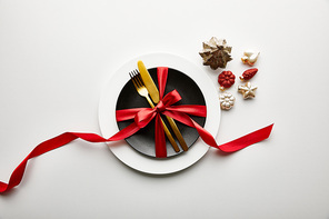 top view of white and black plates with cutlery and ribbon near festive Christmas baubles on white background