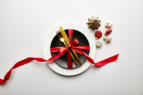 top view of white and black plates with cutlery and ribbon near festive Christmas baubles on white background