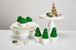 delicious Christmas tree cupcakes with plates and shiny baubles on white surface isolated on grey