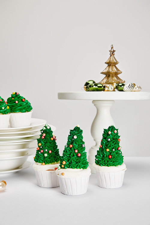 delicious Christmas tree cupcakes with plates and shiny baubles on white surface isolated on grey