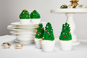delicious Christmas tree cupcakes with plates and shiny baubles on white surface isolated on grey