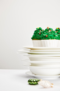 selective focus of delicious Christmas tree cupcakes with plates and shiny baubles on white surface isolated on grey
