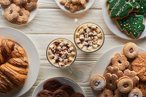 top view of cacao with marshmallow and cacao powder in mugs on white wooden table with Christmas cookies and croissants