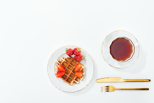 top view of waffle with strawberries on plate near cup with tea, fork and knife on white